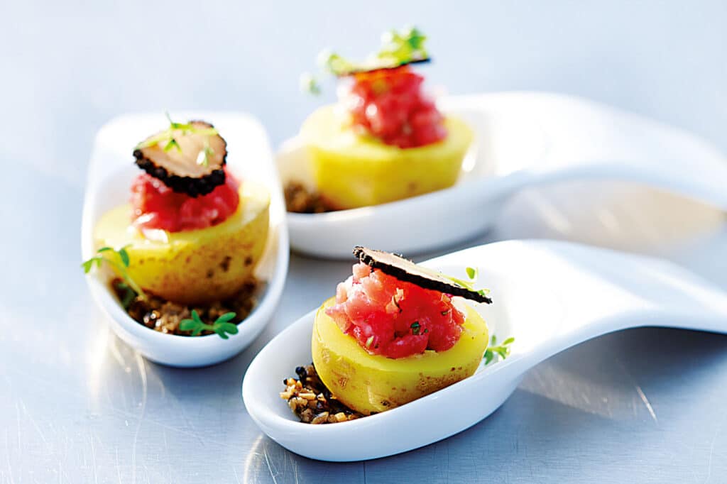 Tartare de Bœuf et Truffe Noire sur Pomme de Terre Amuse-bouche de tartare de bœuf sur pomme de terre, garni de truffe noire et herbes, servi en cuillères de porcelaine.